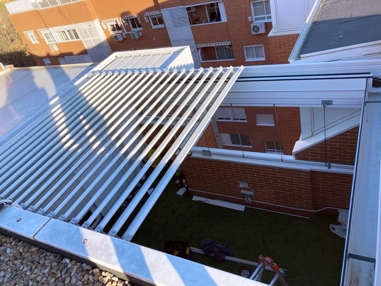 Vista superior de una pérgola con lamas blancas parcialmente abiertas, a través de las cuales se ve el césped