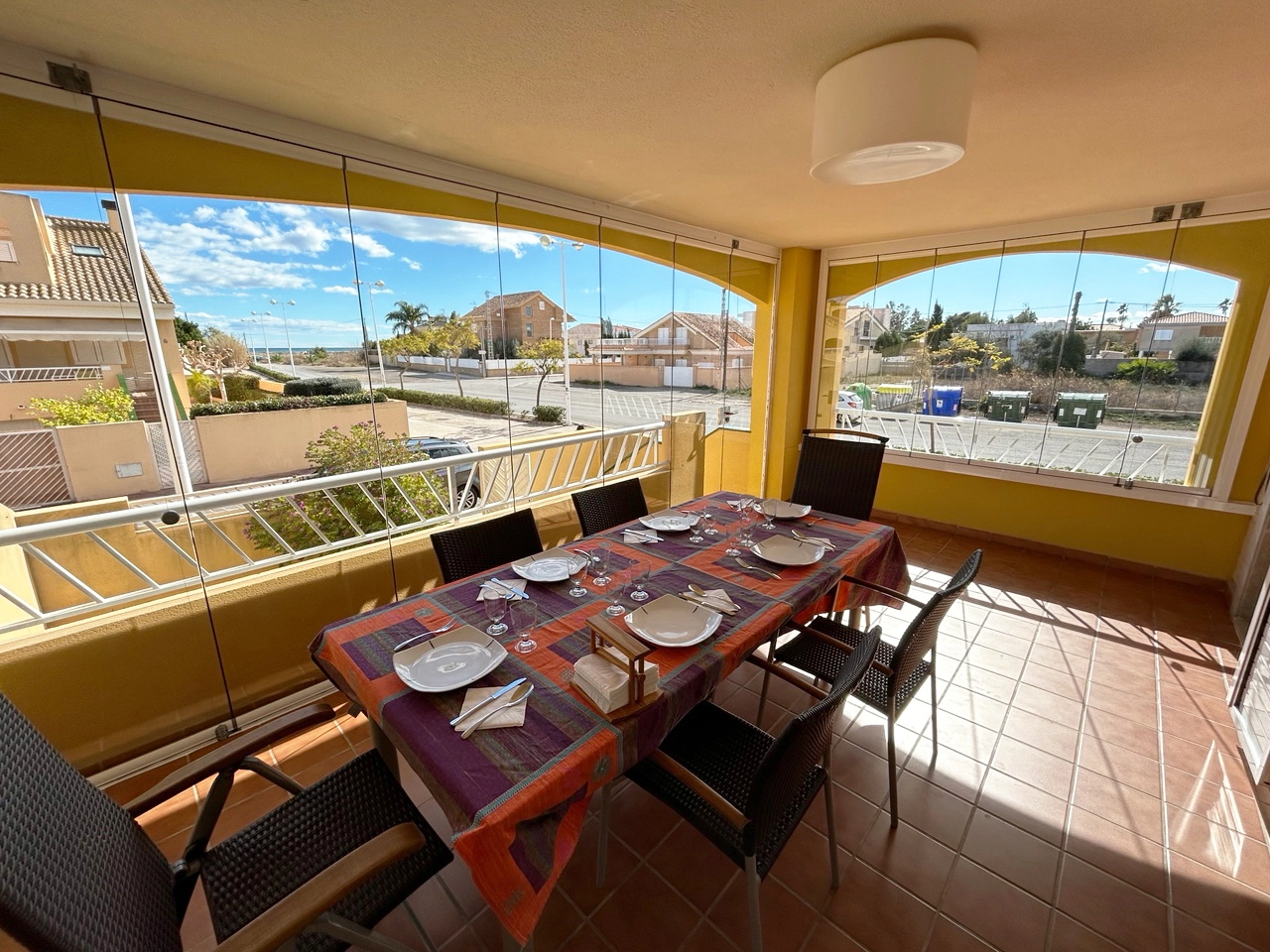 En la foto se muestra una terraza acristalada con una mesa de comedor cubierta con un mantel colorido. En la mesa están colocados platos, cubiertos y otros utensilios.