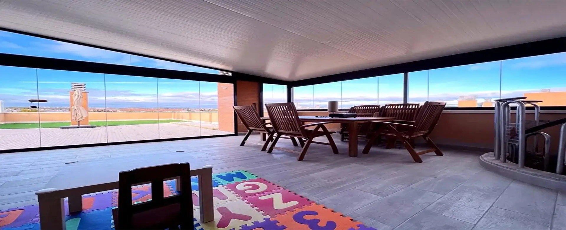 Una veranda modernizada con acristalamiento panorámico, muebles de madera y una alfombra infantil en el suelo.