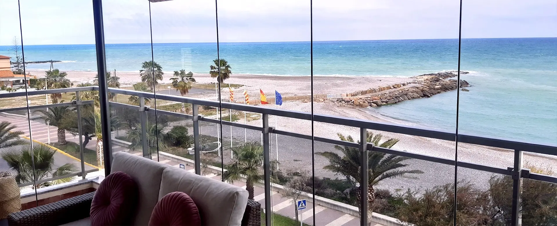Un balcón con acristalamiento panorámico y vistas a la playa, palmeras y el mar, con muebles acogedores en primer plano.